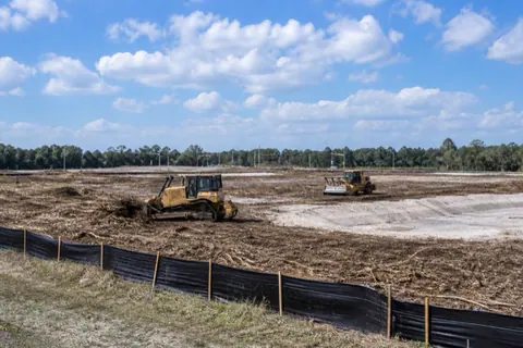 Pair of unbranded machines opening a large commercial parcel along a Gainesville arterial, windrowed mulch and perimeter silt fence in place
