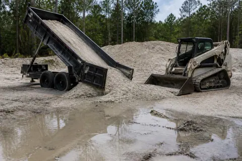 Pale fill sand pouring from a dump trailer at a soggy lot corner as a compact loader pushes it level