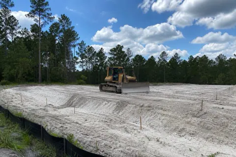 Compact dozer leveling pale sand across a staked homesite pad with silt fence along the low edge