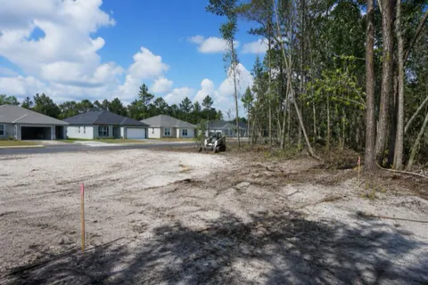 Half-cleared quarter-acre lot with pale sand exposed, surveyor stakes flagged pink, and new homes across the street