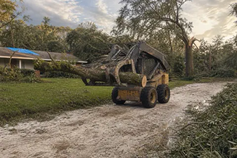 Snapped water oak trunk lifted by grapple at the edge of a storm-littered Gainesville yard, limb piles staged